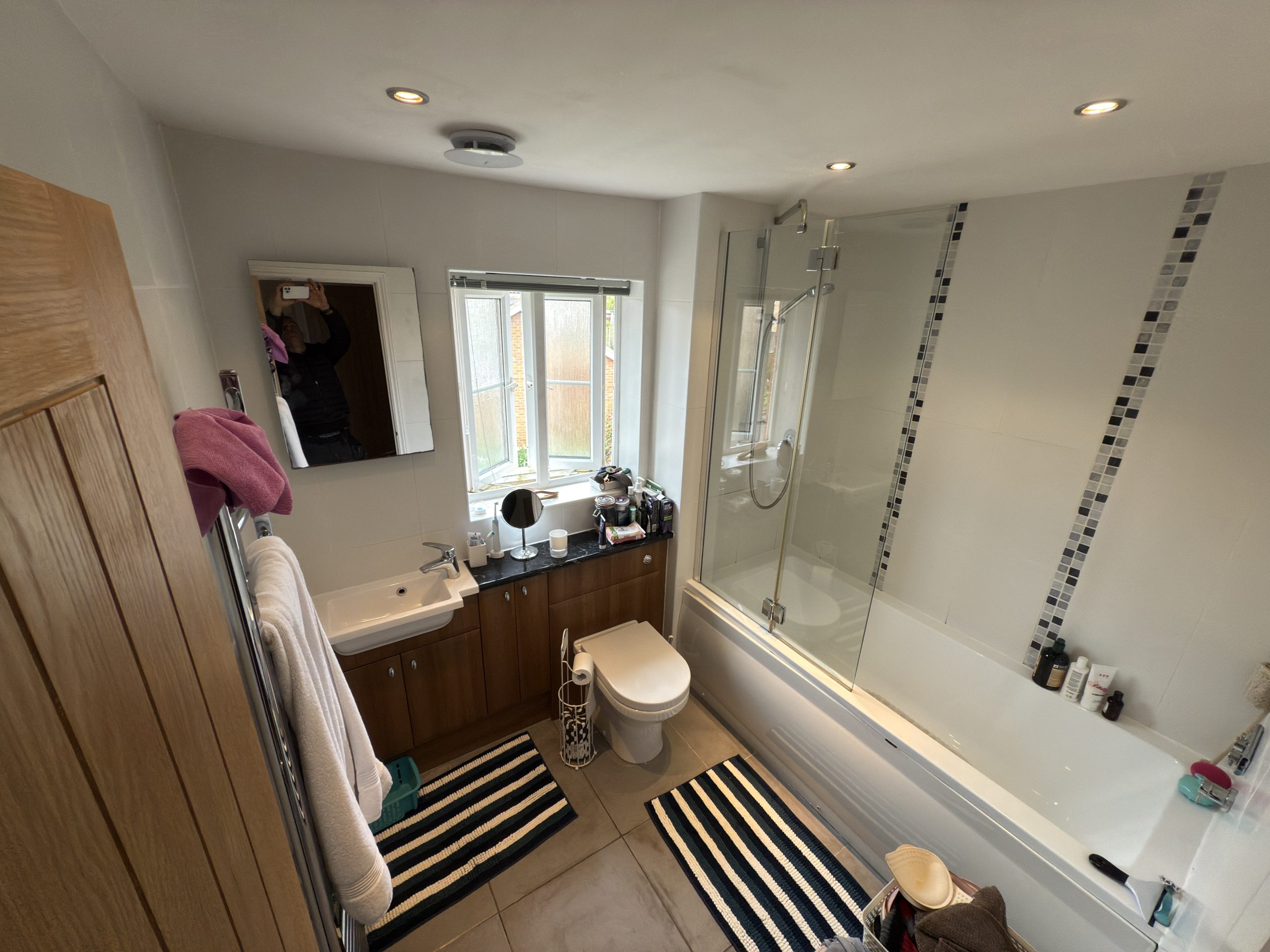 Modern bathroom with a bath-shower combo, toilet, sink with mirror, and striped rugs. Kitchen and bathroom showroom design inspiration is reflected in the layout. Toiletries sit on the worktop while natural light streams through two windows.
