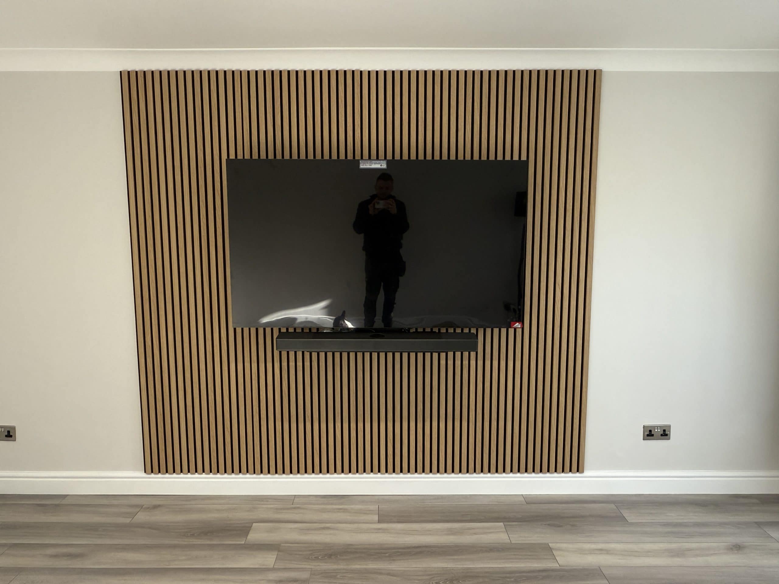 A flat-screen TV is mounted on a wall with vertical wooden slats in a modern kitchen and bathroom showroom. Below the TV is a slim black shelf. The light wood floor adds warmth, and a person’s reflection appears in the screen.