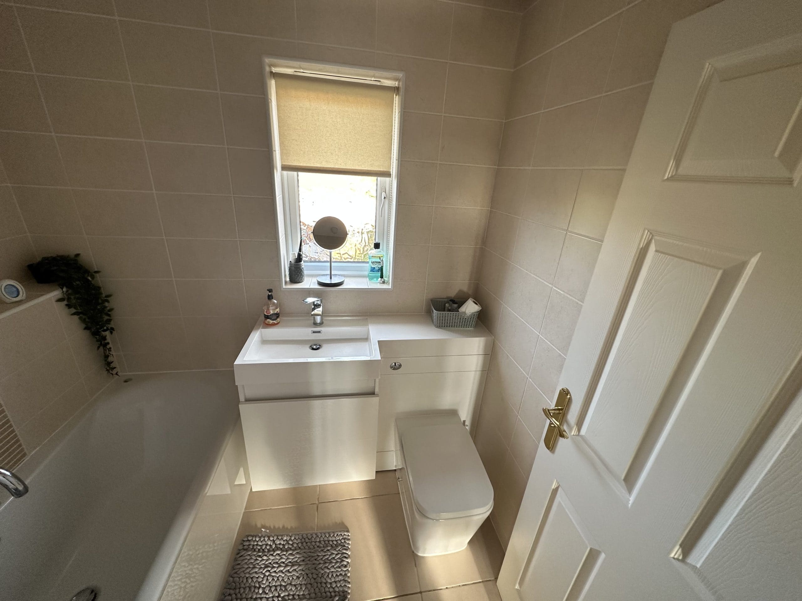 A small beige-tiled bathroom, designed with showroom-inspired touches, features a bath to the left, white vanity and basin, mirror, soap and basket, toilet, shaded window, and a partly open door—highlighting quality installation and supply details.