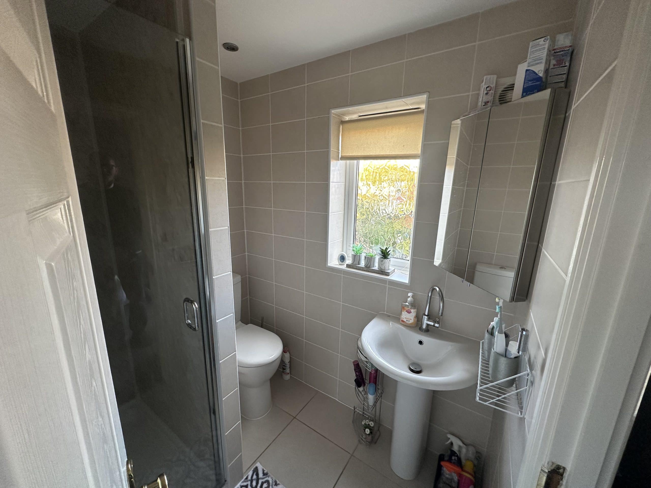 A small bathroom with beige tiled walls, a glass shower door, a white toilet, and a pedestal sink showcases thoughtful bathroom design. Several toiletries and plants on the windowsill add charm—perfect inspiration for any kitchen and bathroom showroom.