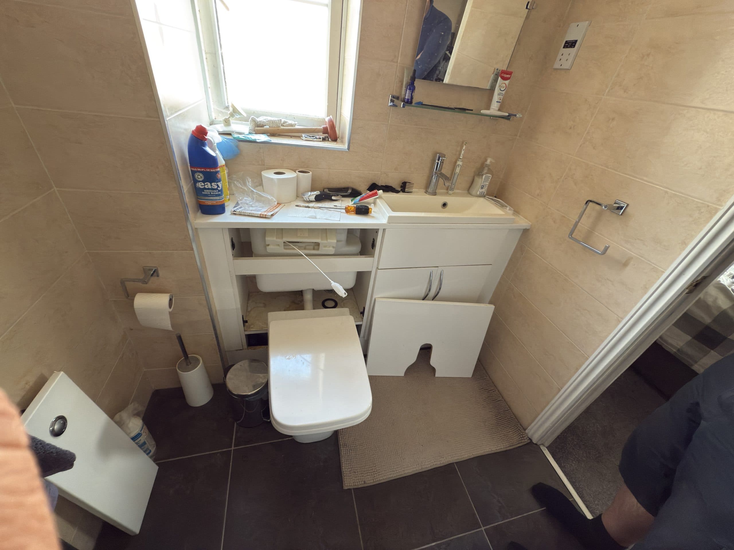 A small beige-tiled bathroom showcases a practical design, with open cupboard doors under the sink and cleaning supplies scattered about—echoing the everyday realities behind kitchen and bathroom showroom installation. A person’s legs are partially visible.