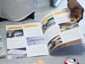 A person holds open a brochure featuring kitchen and bathroom showroom design ideas and installation supplies on a table. A fizzy drink can sits nearby, while the pages display photos and descriptions in yellow and white sections.