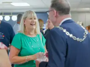 A woman in a green top smiles and chats with a man in a suit and ceremonial chain at a social event in a brightly lit room, possibly discussing kitchen and bathroom showroom design. Other people are visible in the background.