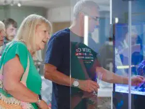 A woman and a man look at a display case in an indoor setting, appearing interested in the kitchen and bathroom showroom installation supplies and designs showcased inside. The man wears a dark shirt and glasses; the woman has blonde hair and a green top.