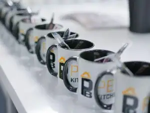 A row of white mugs with black handles and spoons inside, lined up on a white worktop in a kitchen and bathroom showroom. The mugs have partial yellow and black lettering that reads “KITCH.”.