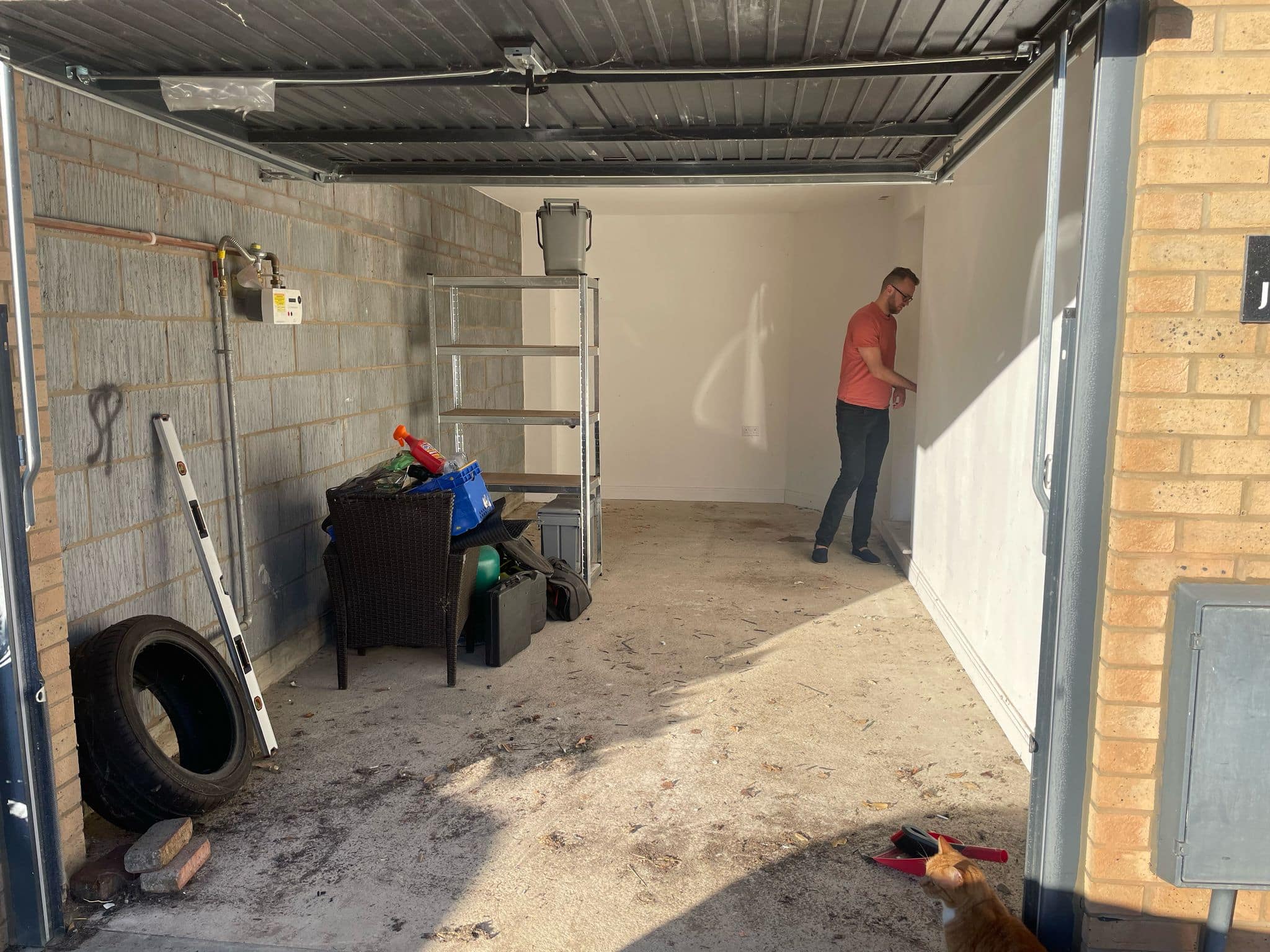 A man in a red shirt stands at the back of a mostly empty garage, with shelves, a tyre, and garden tools on the left. A cat sits near the entrance in sunlight. The space hints at potential for kitchen and bathroom showroom design or installation supply storage.
