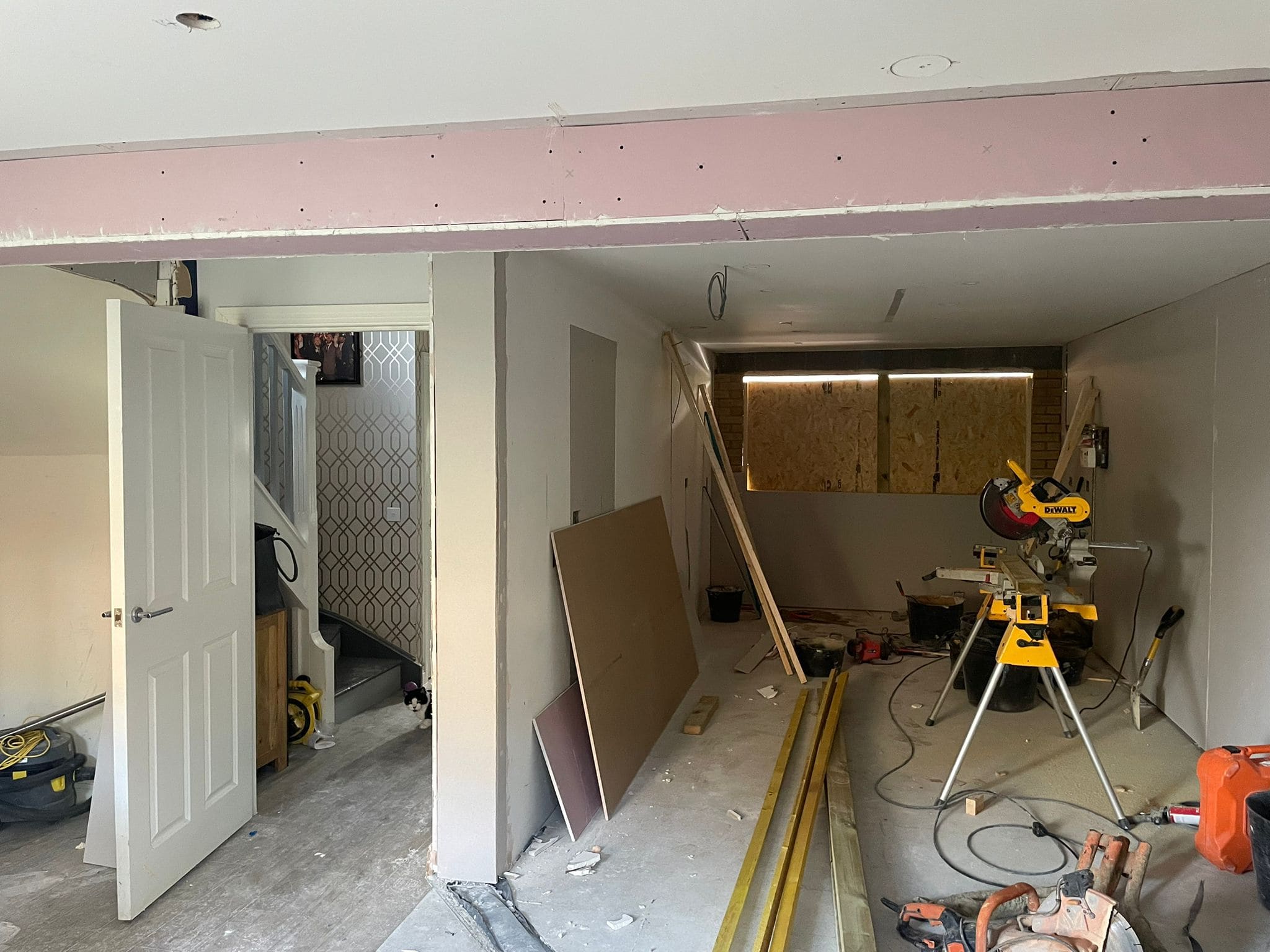 A room undergoing renovation with construction tools, boards, and materials scattered on the floor. An open door leads to a staircase, and a window is boarded up. The space suggests a future kitchen and bathroom showroom installation in progress.