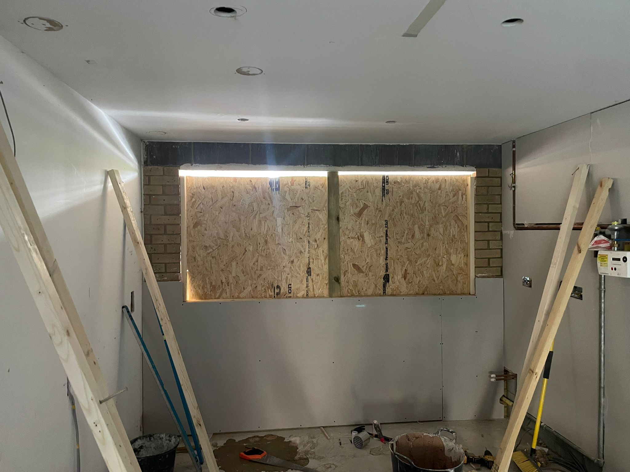 A partially renovated room with exposed plywood covering a window, brickwork on either side, tools and wooden supports around, and unfinished walls and ceiling—ideal for kitchen and bathroom showroom installation or design. Construction materials are scattered on the floor.
