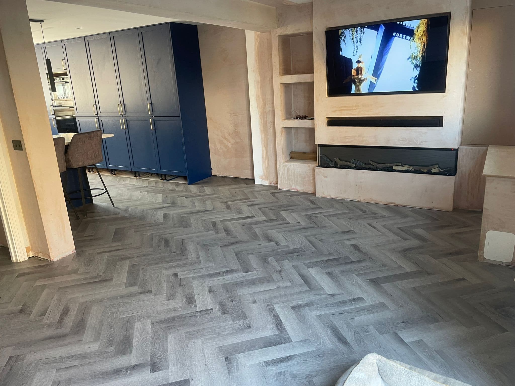 A modern living room with light grey herringbone flooring, a wall-mounted TV showing a person, a built-in fireplace, light walls, and blue kitchen cupboards—perfect inspiration for any kitchen and bathroom showroom design.
