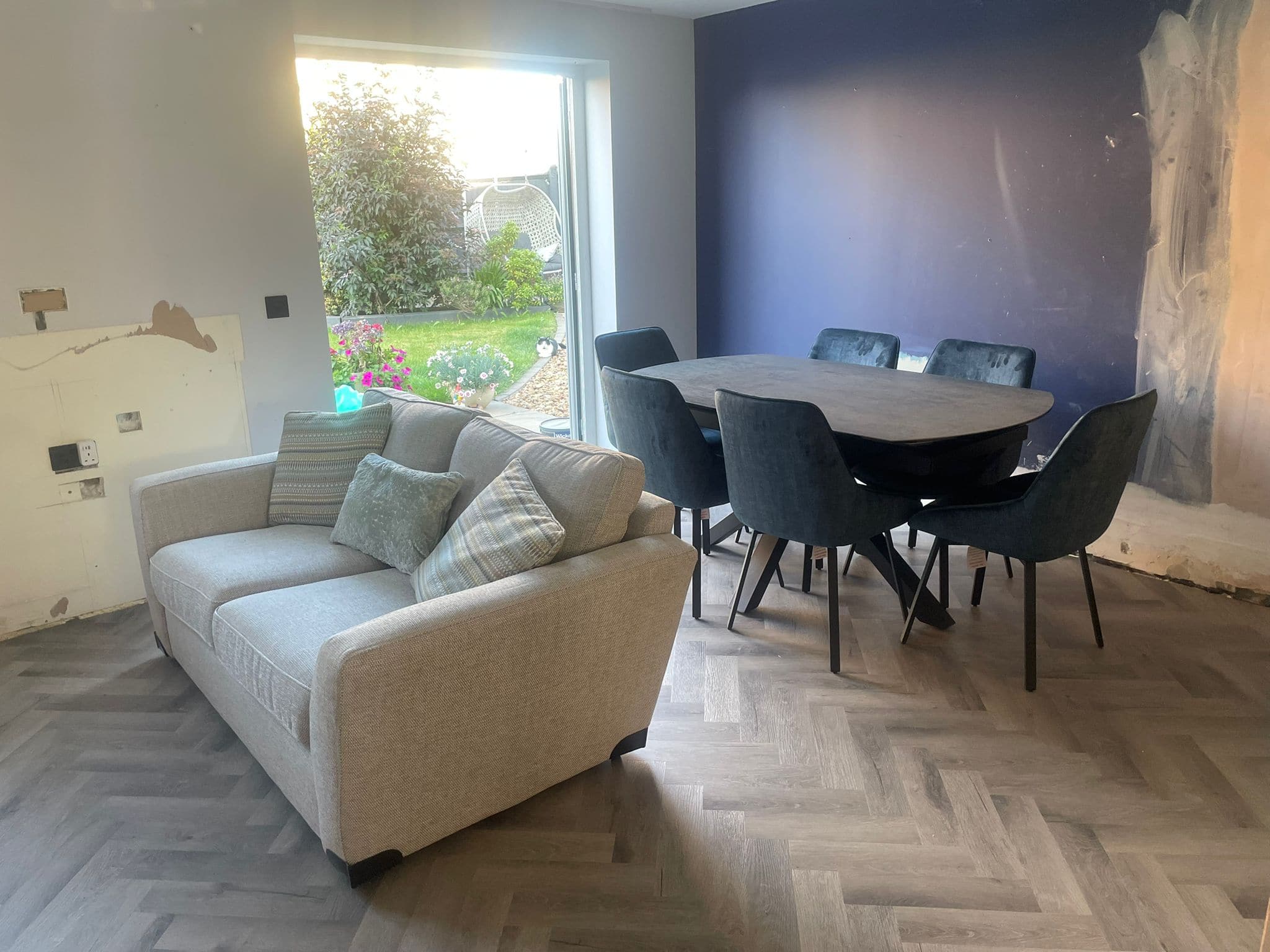 A modern room with a light grey sofa and cushions, a dark oval dining table, wood-patterned floor, and a large window overlooking the garden—perfect inspiration for your next kitchen and bathroom showroom installation or design project.