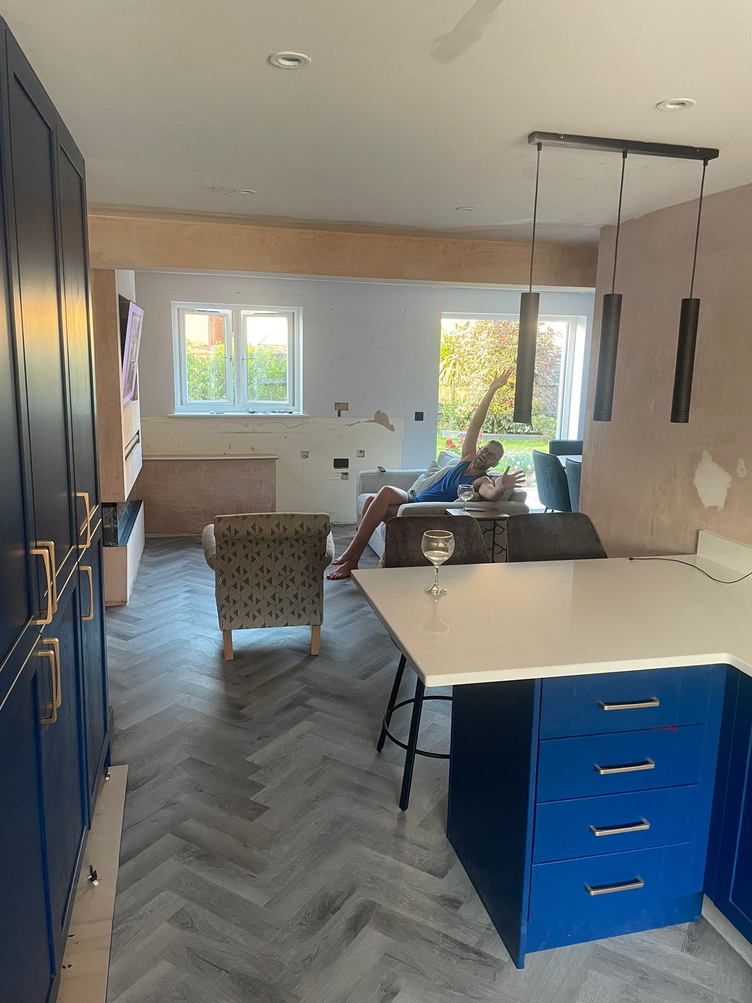 A person relaxes on a sofa in a partially renovated kitchen and living area with unfinished walls, blue cupboards from a kitchen and bathroom showroom, herringbone flooring, pendant lights, and a glass of wine on the white worktop.