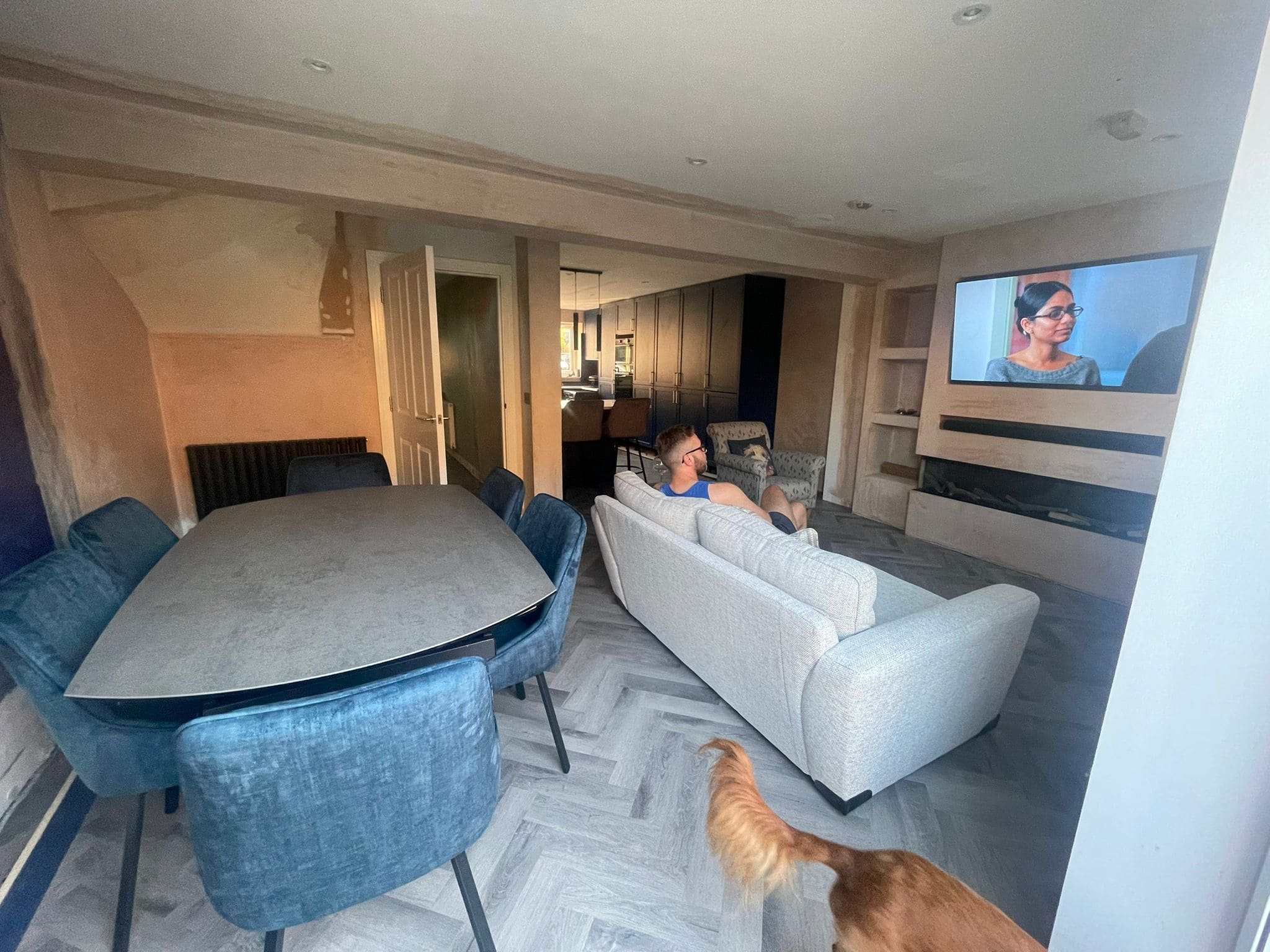 A man sits on a light-coloured sofa watching TV in a modern open-plan living and dining area, showcasing sleek design. A large dining table with blue chairs is nearby, hinting at inspiration from kitchen and bathroom showroom installations. A dog’s tail is visible in the foreground.