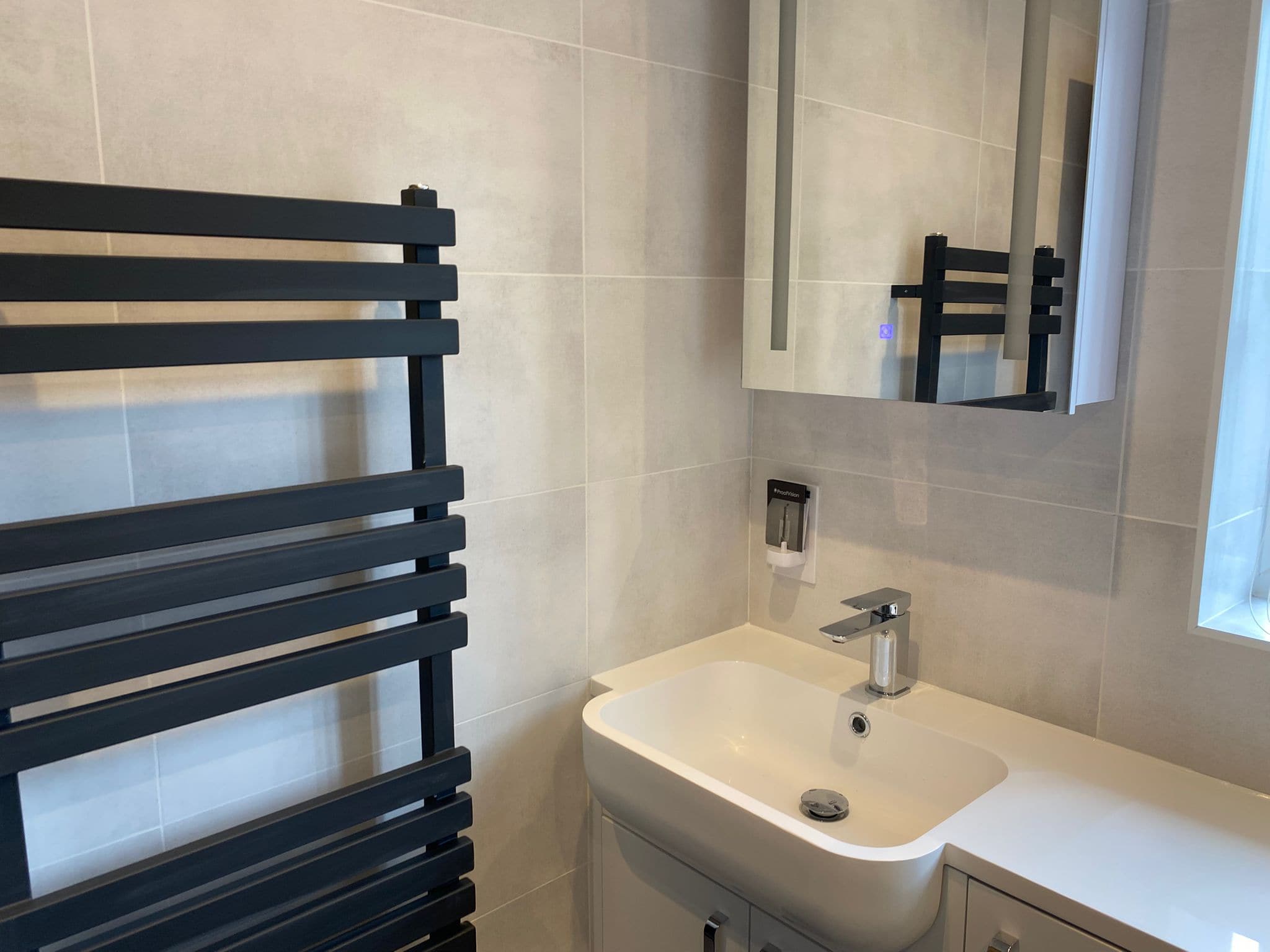Modern bathroom with light grey tiled walls, a black heated towel rail, a white sink with chrome tap, and natural light—showcasing expert bathroom design perfect for any kitchen and bathroom showroom installation.