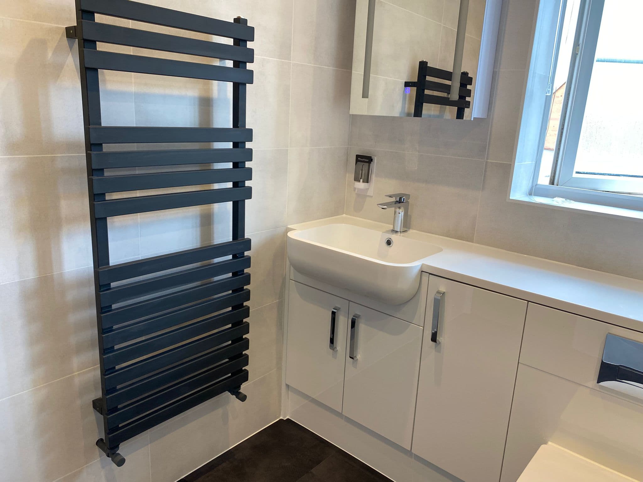 A modern bathroom designed with a black heated towel rail, a white sink with storage cupboard below, mirrored cabinet above, and a window for natural light—perfect inspiration from our kitchen and bathroom showroom.