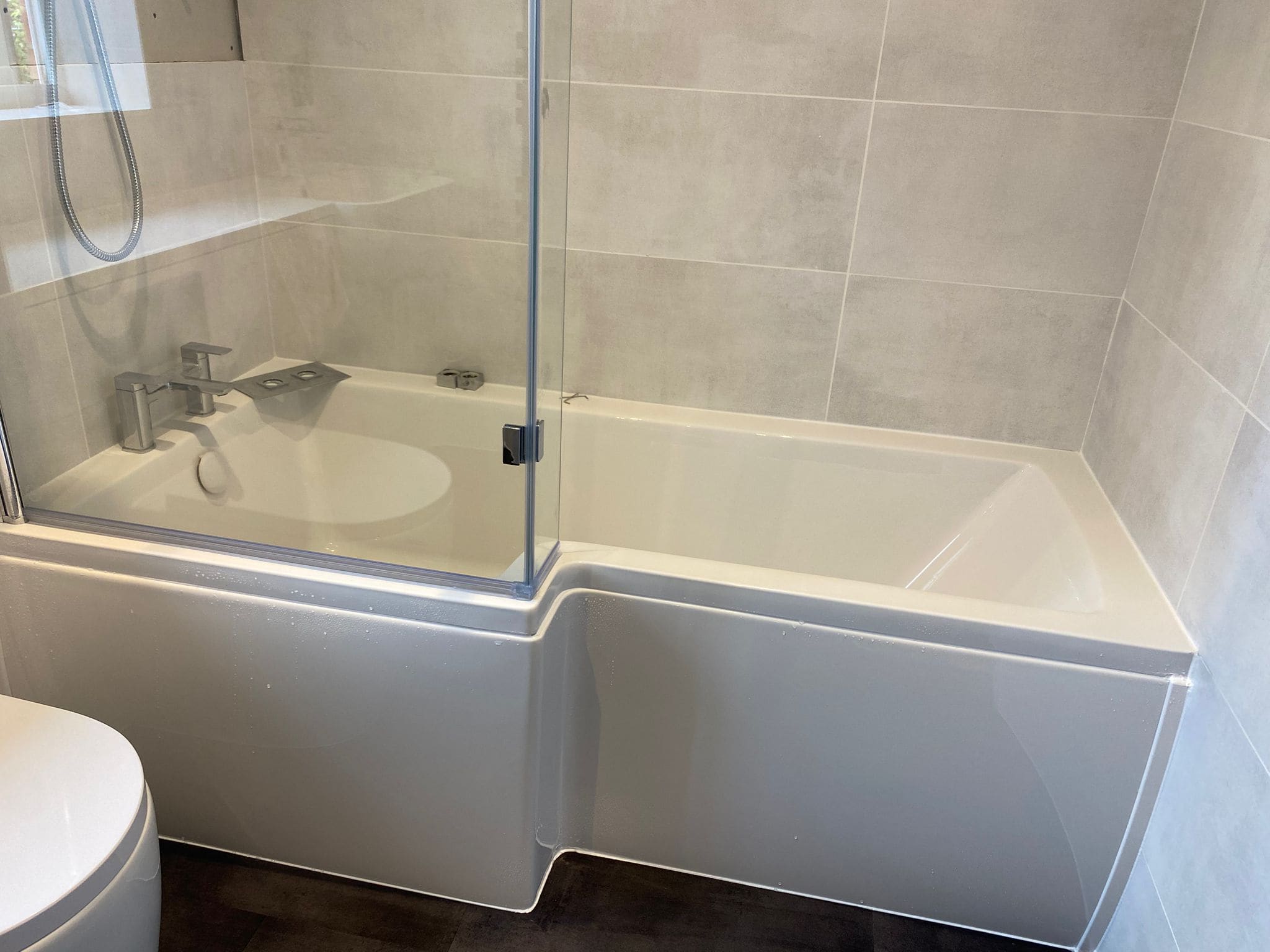 A modern bathroom from a leading kitchen and bathroom showroom, featuring a white bath with a glass shower screen, silver tap, and tiled wall. Part of a white toilet is visible in the lower left corner.