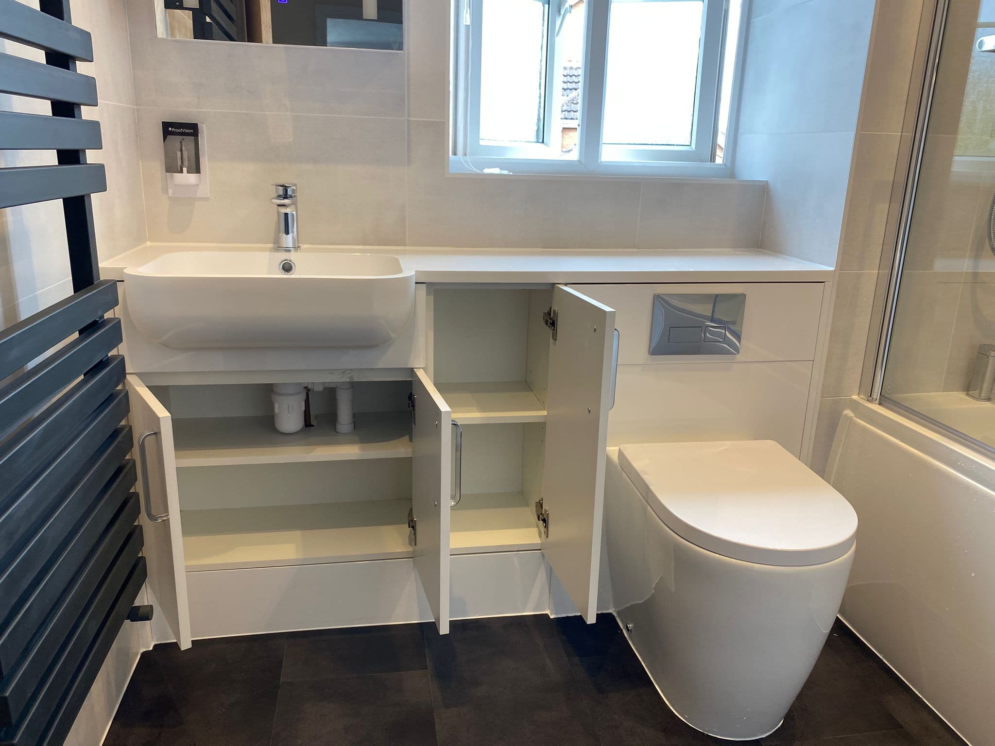 Modern bathroom with a white basin, open cupboard doors revealing shelves and plumbing, a toilet, wall-mounted soap dispenser, window, and part of a bath with glass screen—ideal inspiration for your next kitchen and bathroom showroom design.