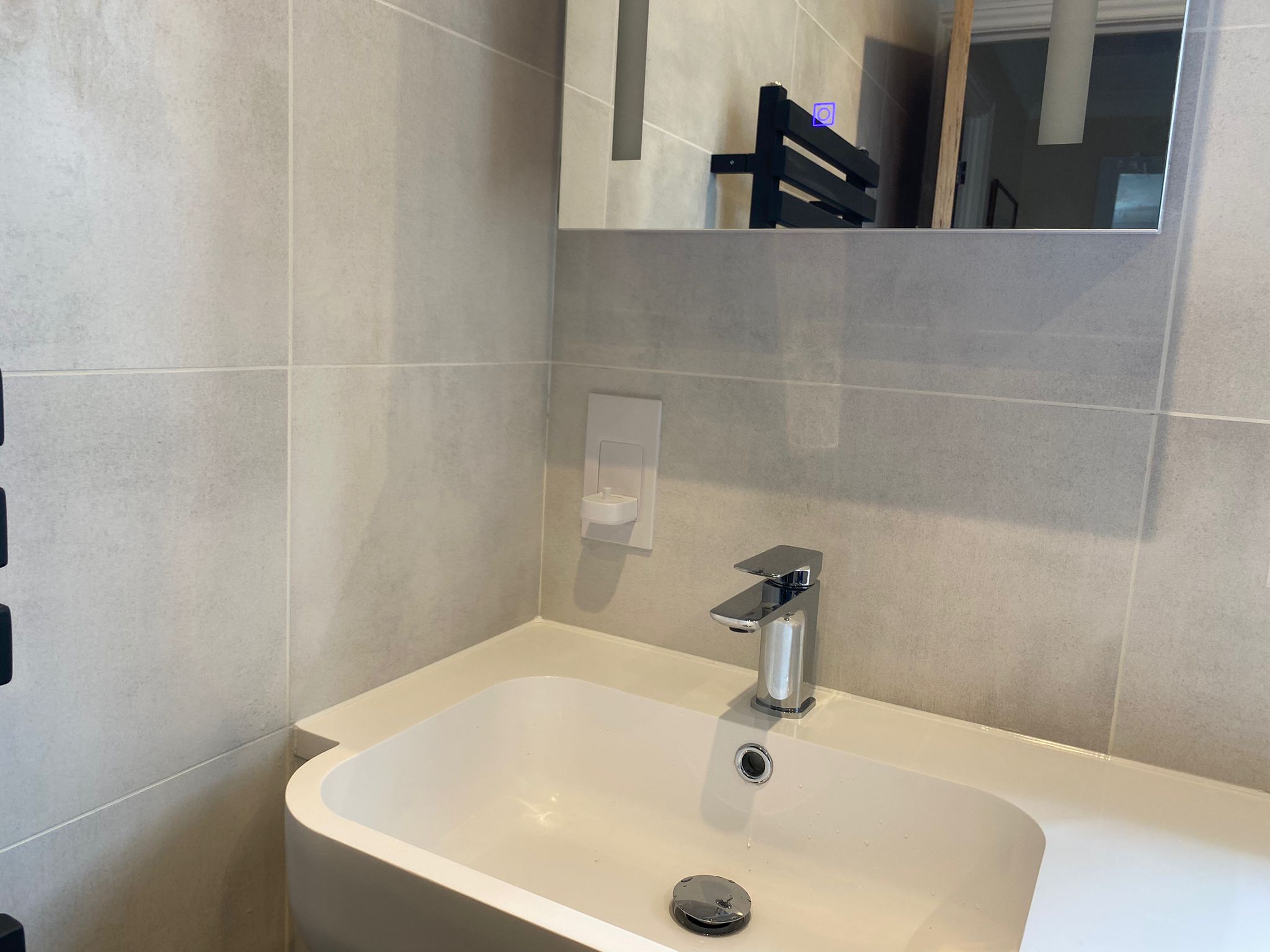 A modern bathroom washbasin from a leading kitchen and bathroom showroom, featuring a rectangular basin, chrome tap, and wall-mounted soap dispenser; a mirror reflects the room and highlights the design with a sleek towel rail.