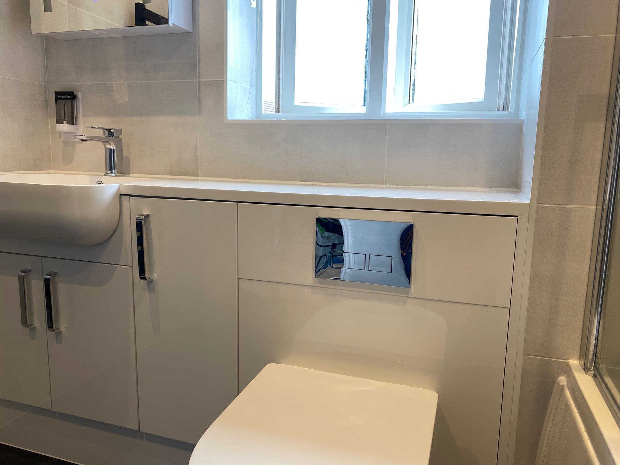 Modern bathroom with white cabinets, a square basin, and a wall-hung toilet beneath a flush plate. A window lets in natural light, and the room’s sleek design highlights quality installation and supply from a premier kitchen and bathroom showroom.
