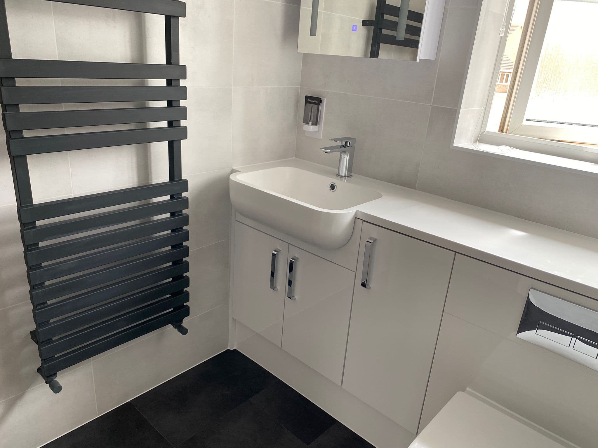 Modern bathroom featuring a black heated towel rail, white sink and cupboards, wall-mounted soap dispenser, and large mirror. Natural light streams through the window onto dark tiled floors—perfect inspiration from a kitchen and bathroom showroom.