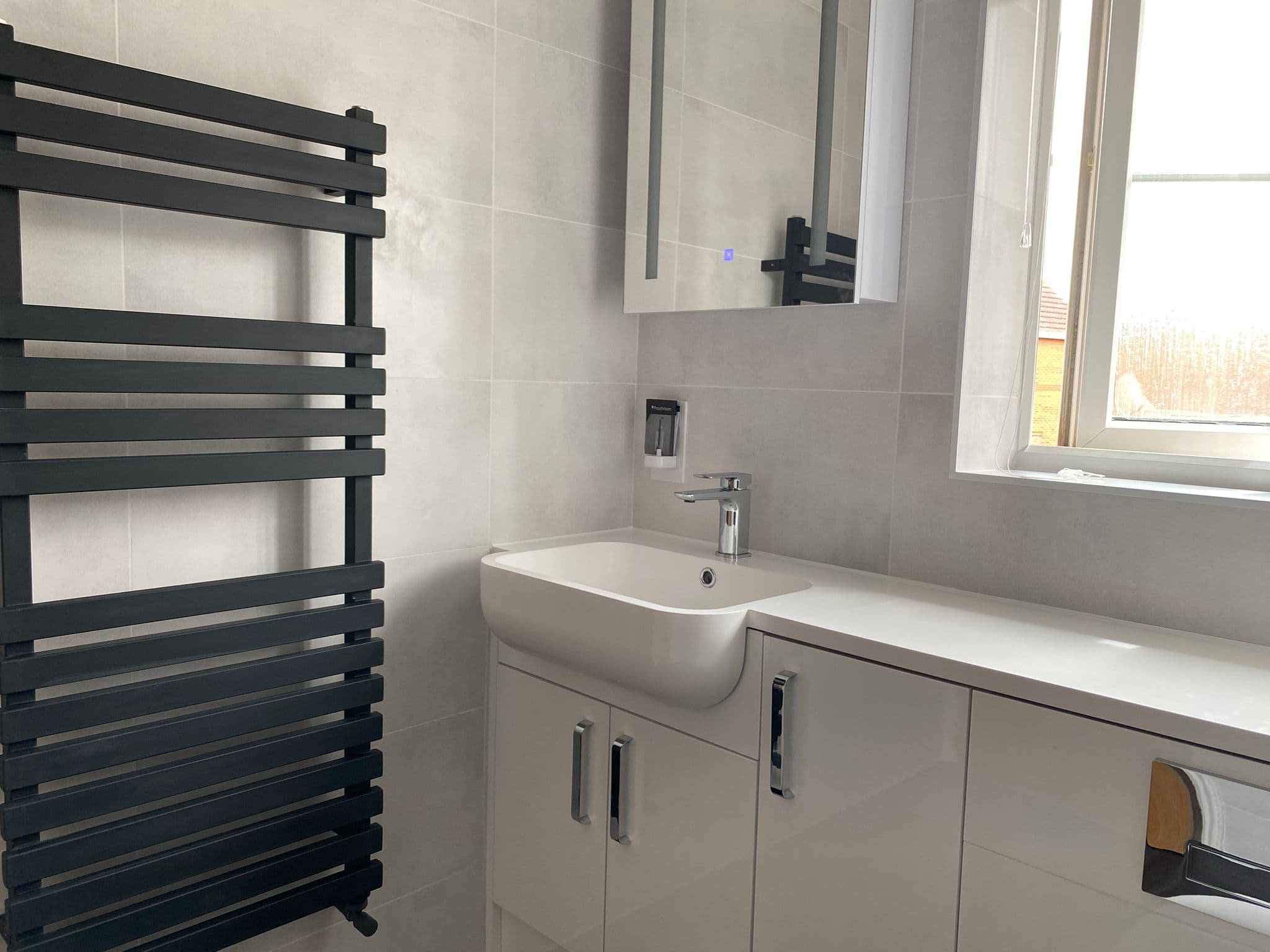 A modern bathroom with light grey tiles, a black heated towel rail, a white sink with a chrome tap, and a mirrored cupboard showcases sleek design—perfect inspiration from a kitchen and bathroom showroom. White cupboards provide storage below the sink.