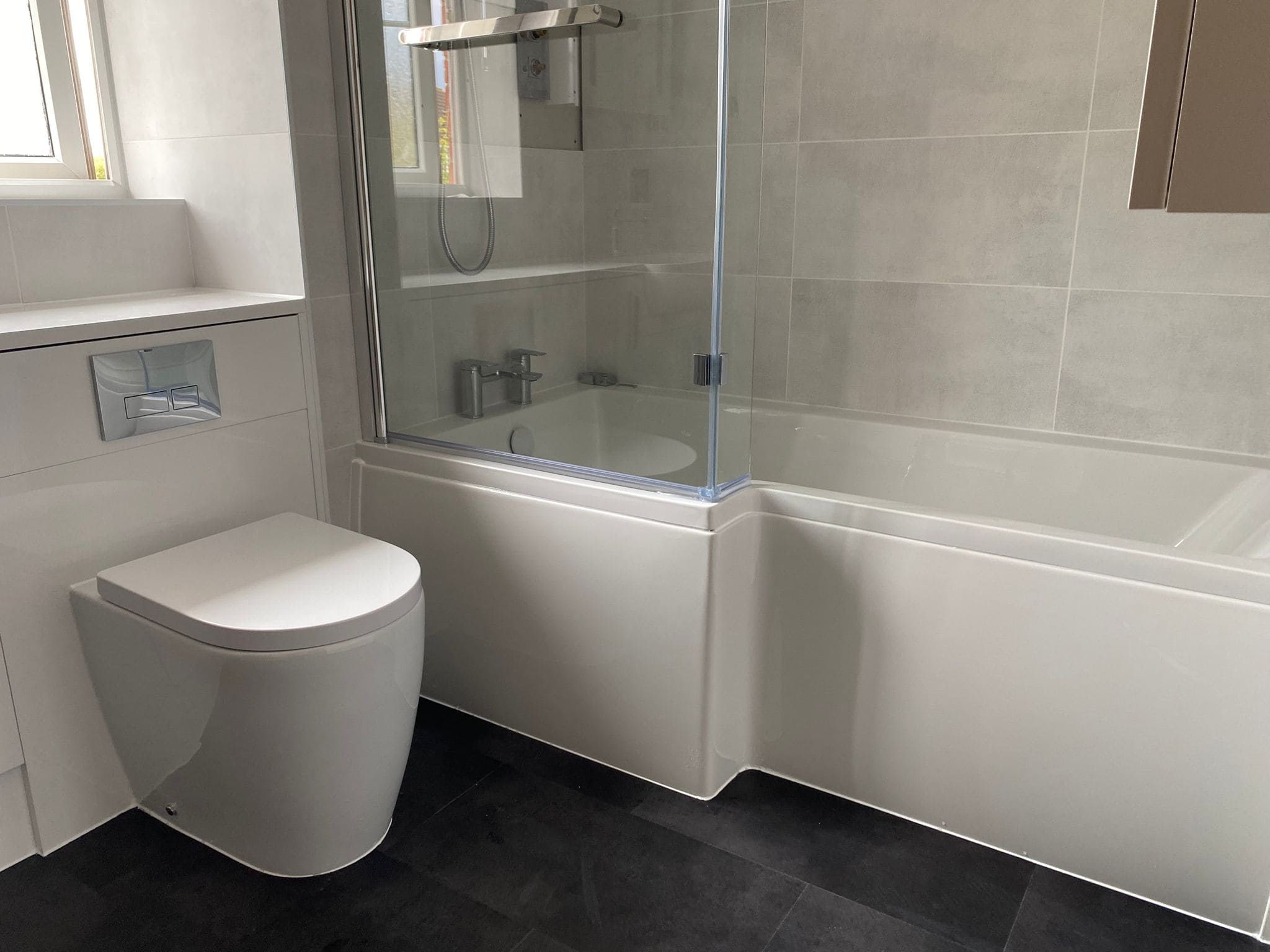 Modern bathroom with a white toilet, a bath-shower combination with clear glass screen, light grey tiled walls, dark floor tiles, and a window letting in natural light—perfect inspiration for any kitchen and bathroom showroom design.