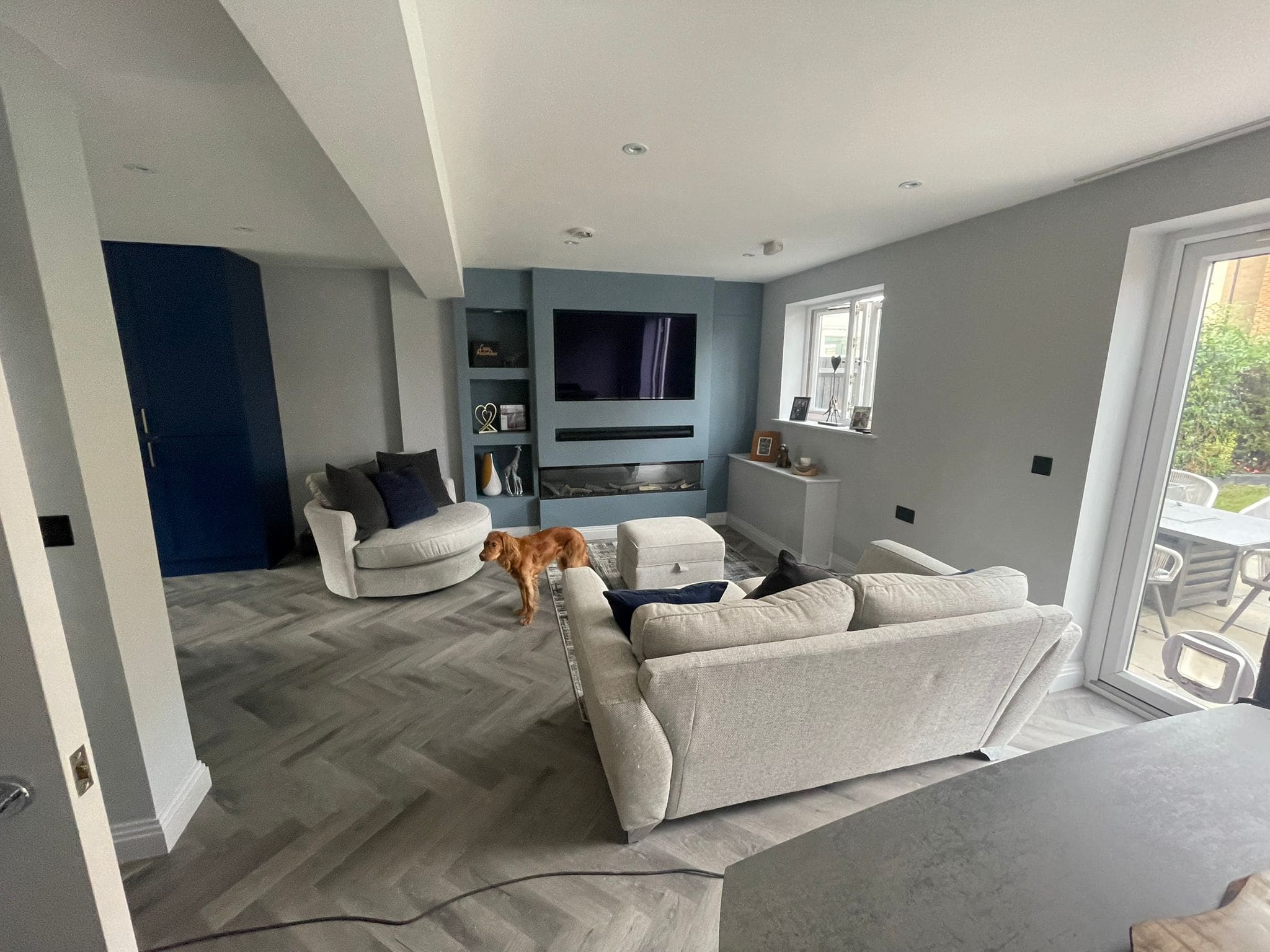 A modern living room with light grey walls and herringbone flooring features two light grey sofas, a round chair, and design elements inspired by kitchen and bathroom showroom installation. A brown dog stands near the seating area as natural light pours in.
