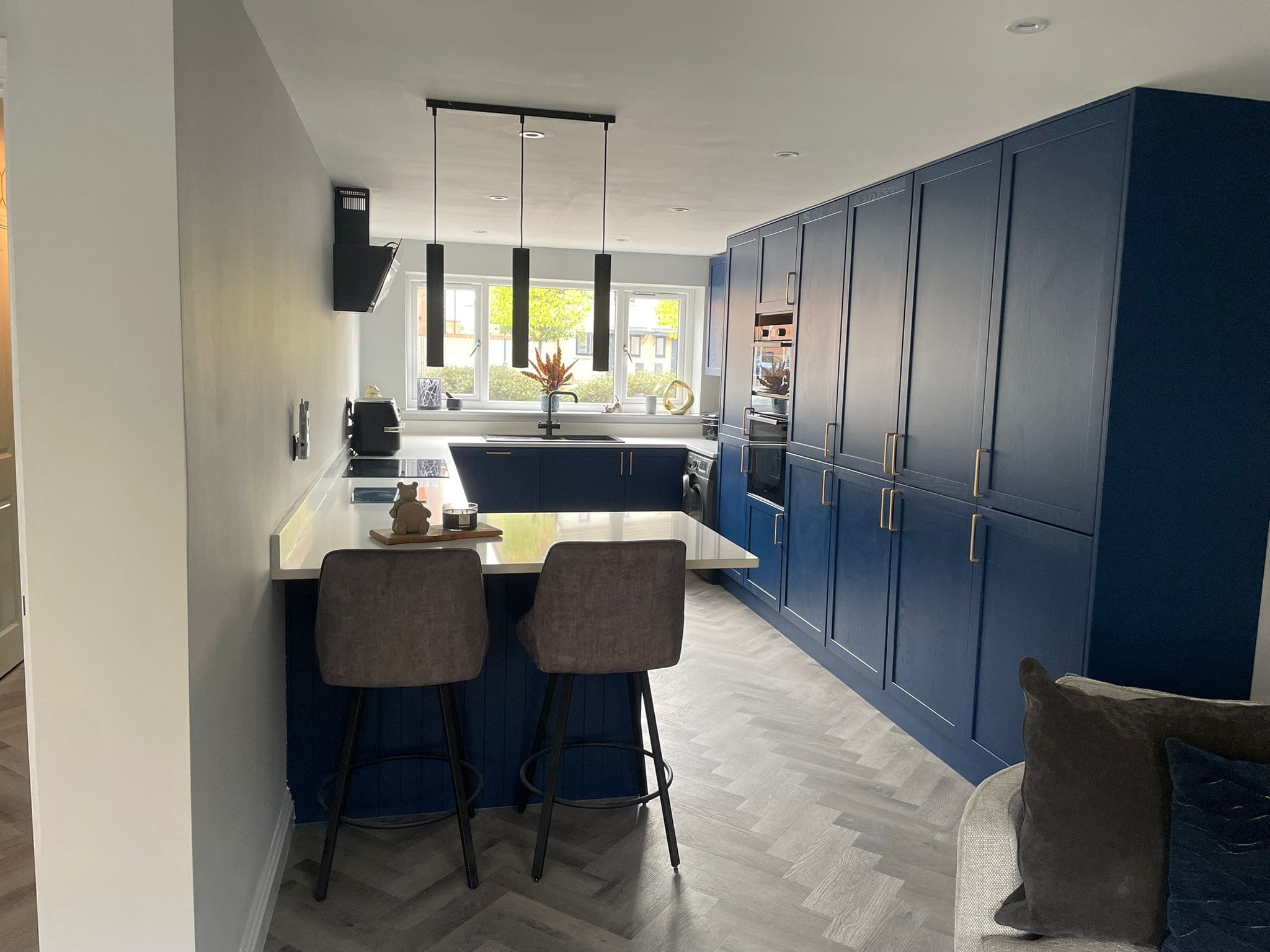 Modern kitchen and bathroom showroom design with blue cabinets, a white worktop, and two grey barstools at the island. Built-in appliances and large windows provide natural light, while herringbone-patterned flooring adds a stylish touch.