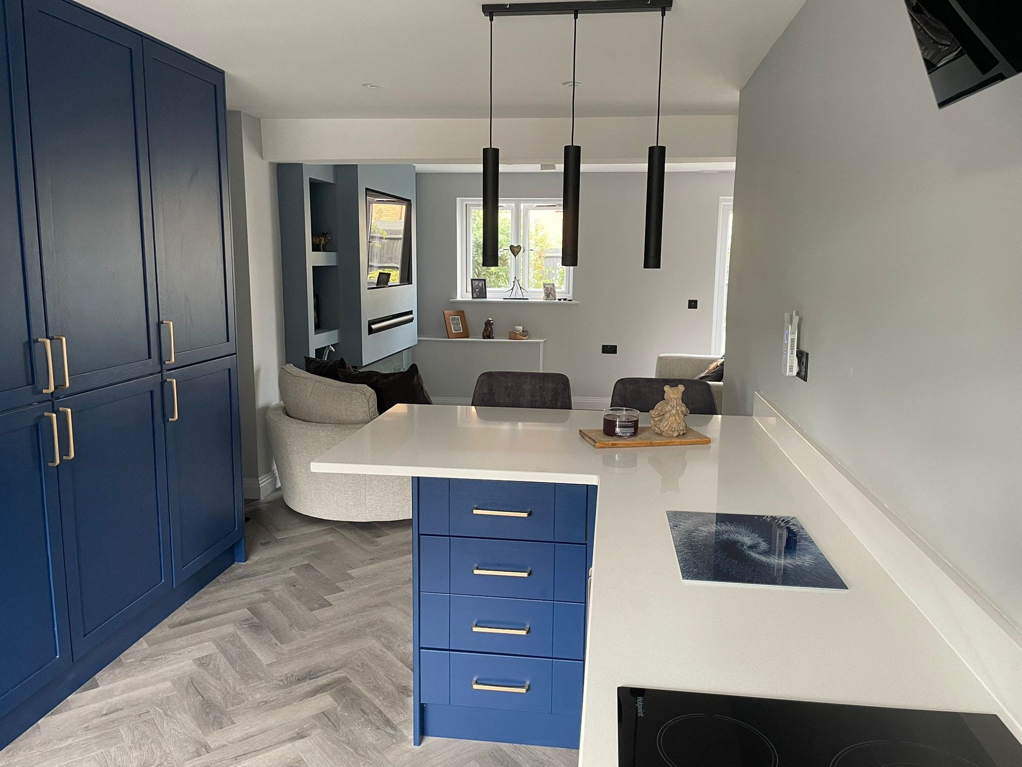 Modern kitchen and bathroom showroom design featuring blue cabinets, white worktops, and a built-in cooker. The island has drawers and pendant lights above, with an open living area and grey sofa visible in the background.