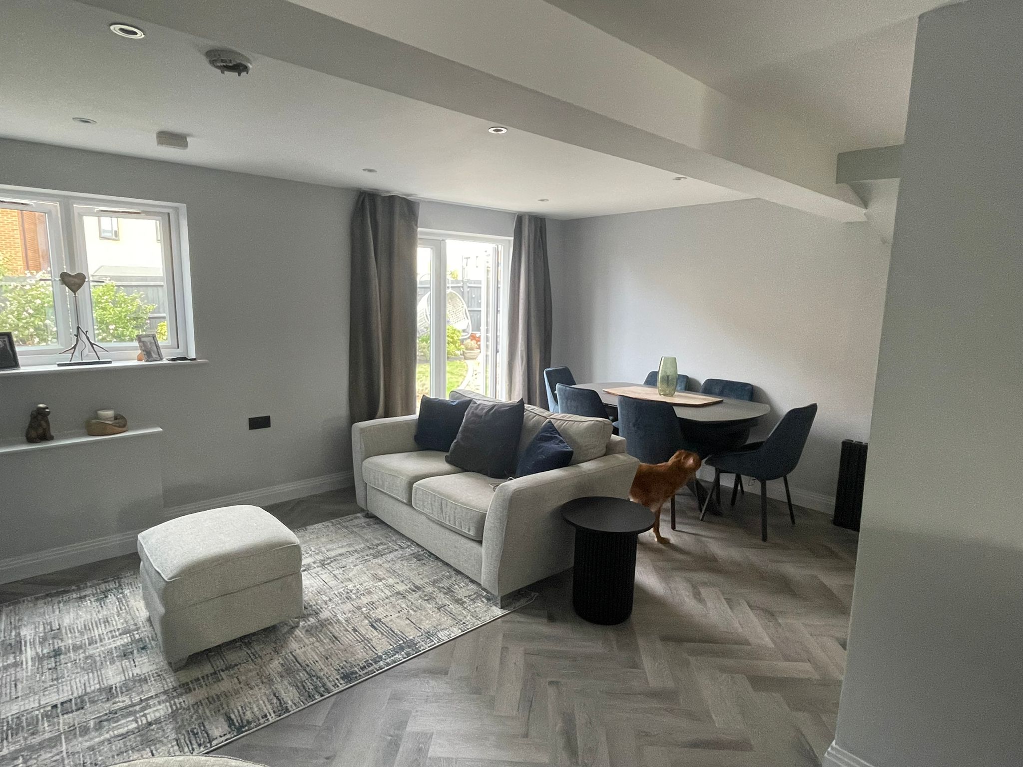 A modern living and dining area with a grey sofa, blue cushions, matching footstool, and a round dining table showcases thoughtful design inspired by kitchen and bathroom showroom installation, with large windows, grey curtains, herringbone floors, and a brown dog.