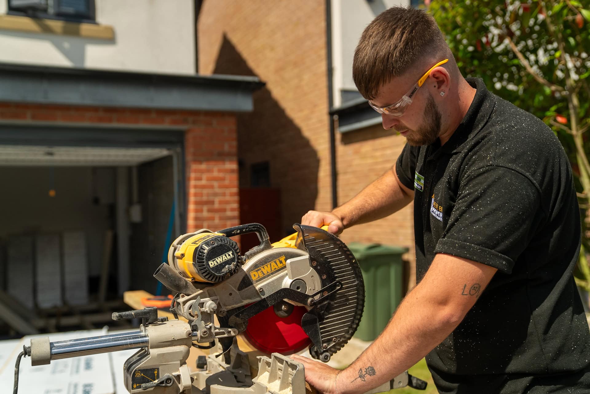 A man wearing safety glasses and ear protection operates a DeWalt mitre saw outdoors, preparing materials for a kitchen and bathroom showroom installation, with a residential garage and greenery in the background.