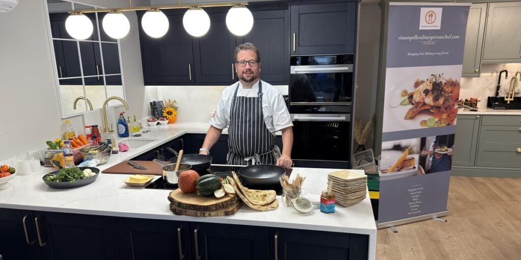 A chef in an apron stands in a modern kitchen behind a counter with fresh vegetables, cooking utensils, and crockery—showcasing the sleek design inspired by a leading kitchen and bathroom showroom installation supplier. Dark cupboards and bright lighting set the scene.