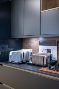 Display of kitchen cupboard samples and laminate finishes arranged on a worktop with dark cabinetry, wood panelling, and modern decor in a showroom setting.