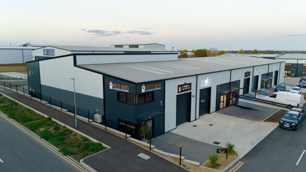 Aerial view of a modern industrial building with multiple units, including Pete Bird Construction, wide entrances, a parking area with vehicles, and surrounding roads during daytime.