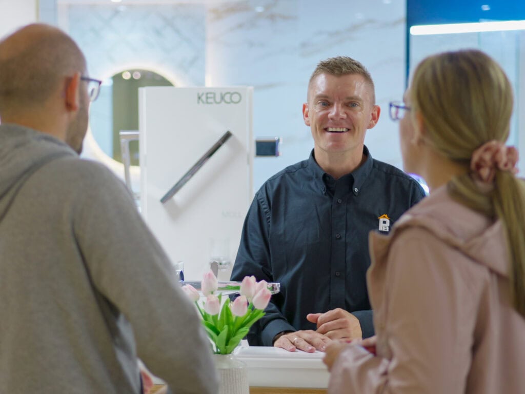 A smiling sales assistant stands at a counter, talking to two customers in a modern showroom.