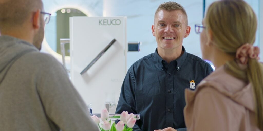 A smiling sales assistant in a black shirt talks to two customers at a counter.