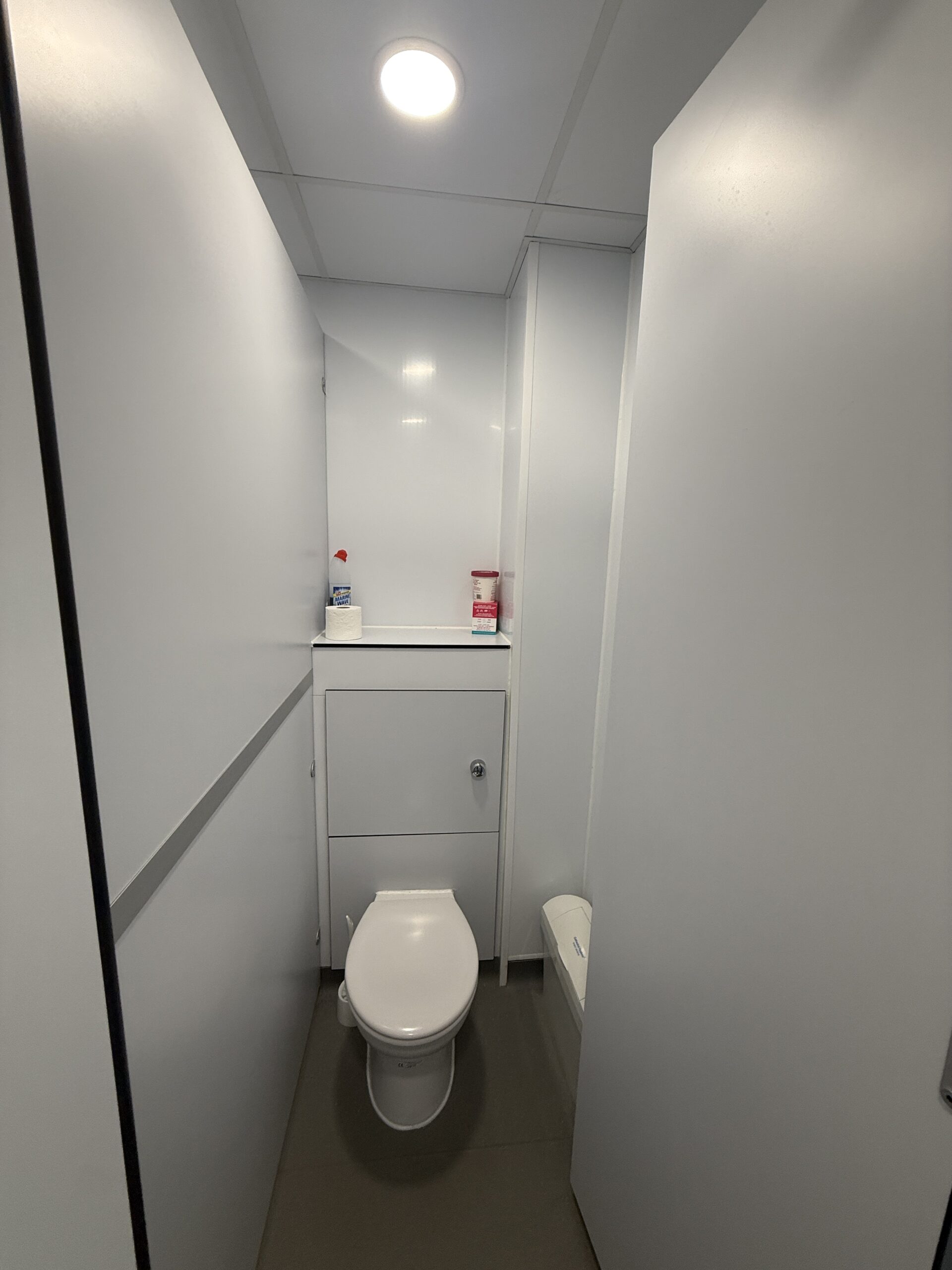 A small toilet with white walls and a toilet. Cleaning supplies and a red-labelled item sit on a shelf above the toilet. Bright ceiling light illuminates the space.