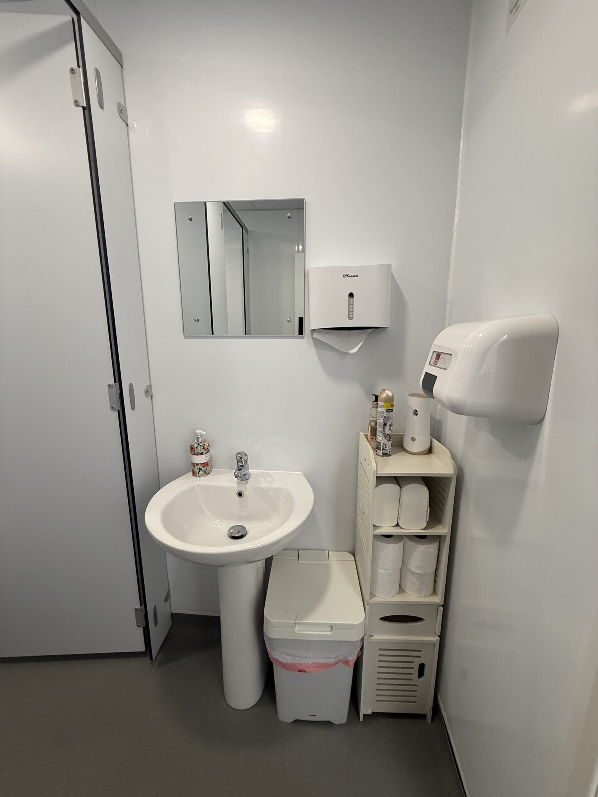 A clean, modern toilet with a white basin, mirror, soap dispenser, hand dryer, rubbish bin, toilet roll holders, and toiletries organised on a small shelf beside the basin. White walls and door add to the minimalist look.