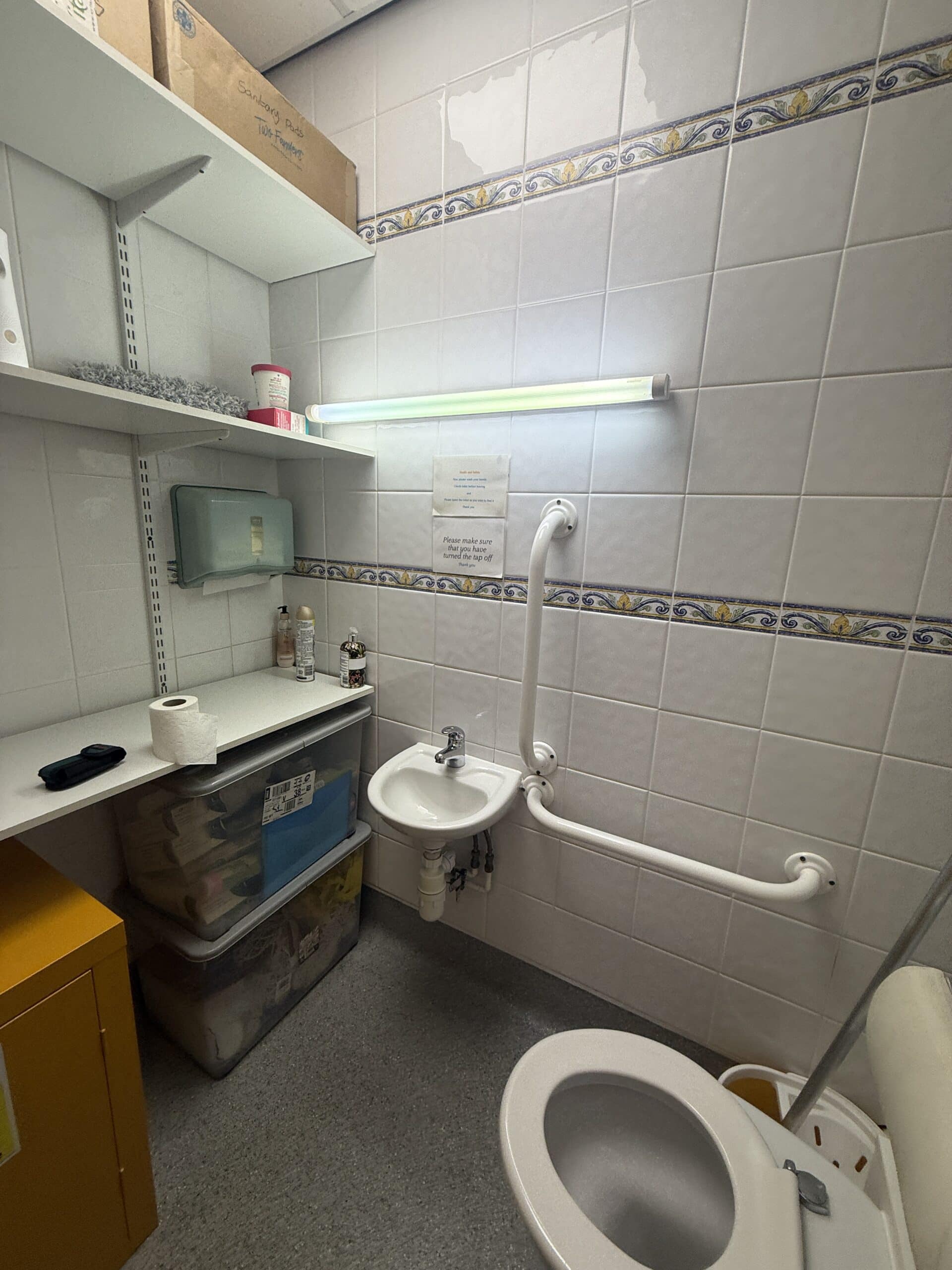 A small toilet with white tiled walls, a toilet with safety rails, a small basin, shelves holding supplies, a fluorescent light above, and storage bins below the shelves. A paper towel dispenser is mounted on the wall.