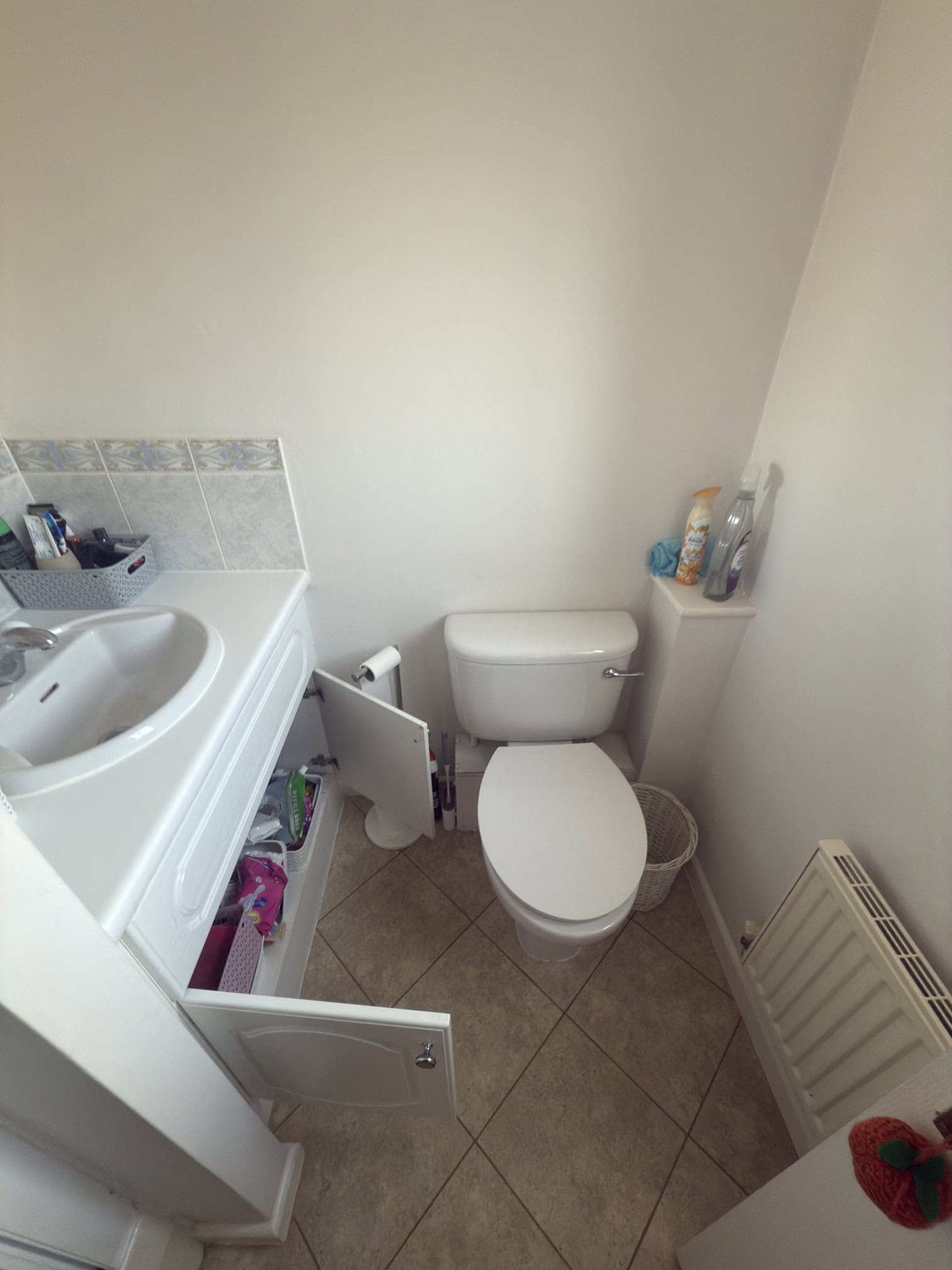 Small bathroom with a washbasin and cupboard, the cupboard doors are open showing toiletries inside. A toilet with a bin beside it, toiletries on top of the toilet cistern, and a radiator on the right wall. Light tiled floor.
