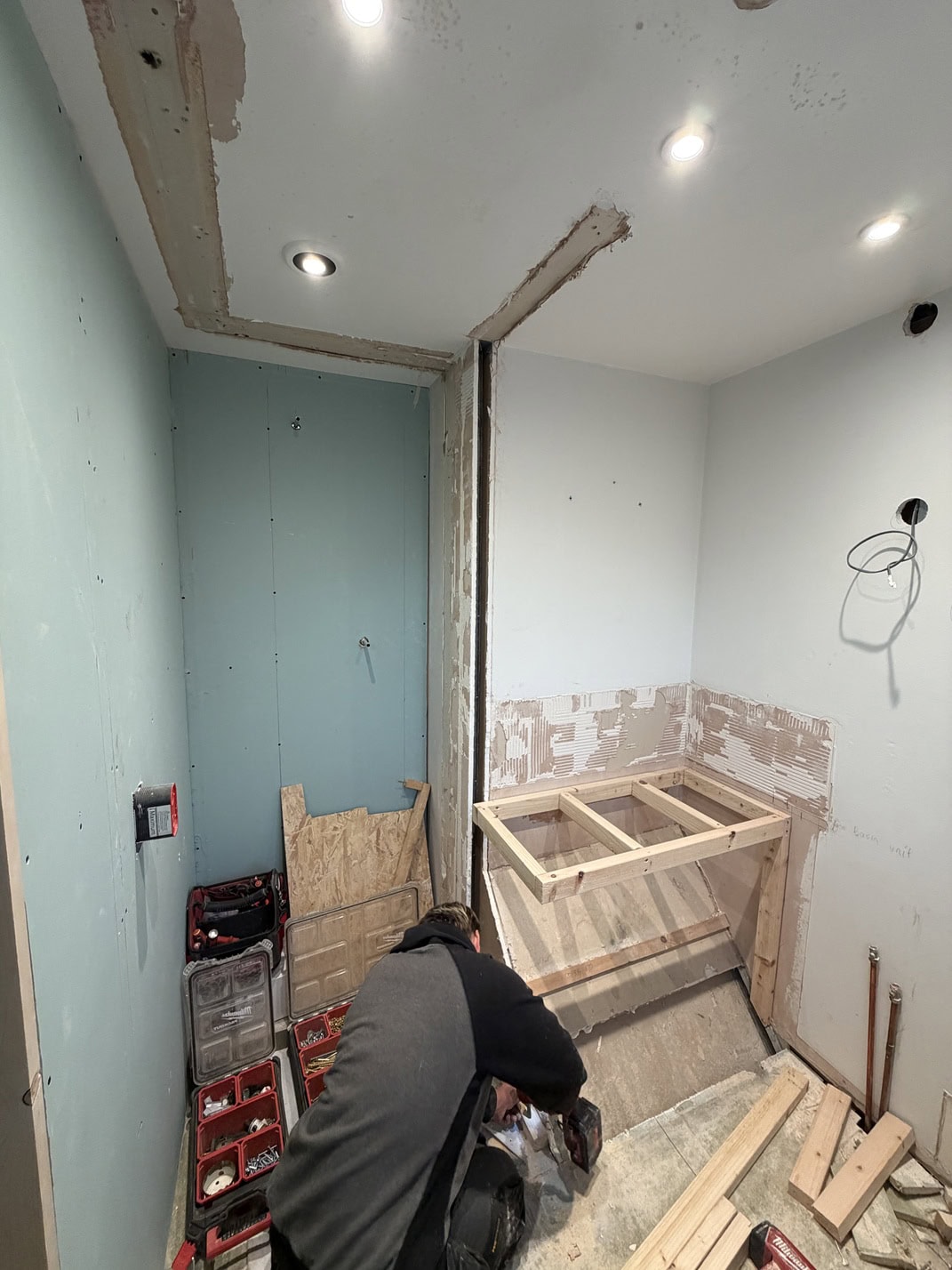 A person is kneeling on the floor of a partially renovated room with tools and materials scattered about. Unfinished wooden framework and exposed plasterboard indicate ongoing construction or refurbishment work.