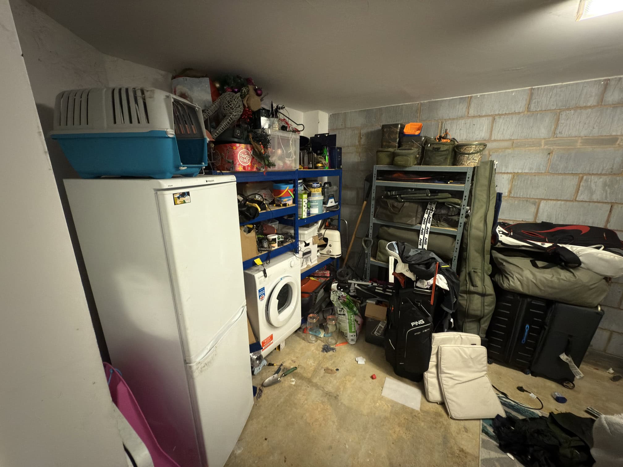 A cluttered utility room with a white fridge, washing machine, blue shelving units filled with various items, bags, and boxes. The floor is scattered with bags, bottles, clothes, and miscellaneous objects.