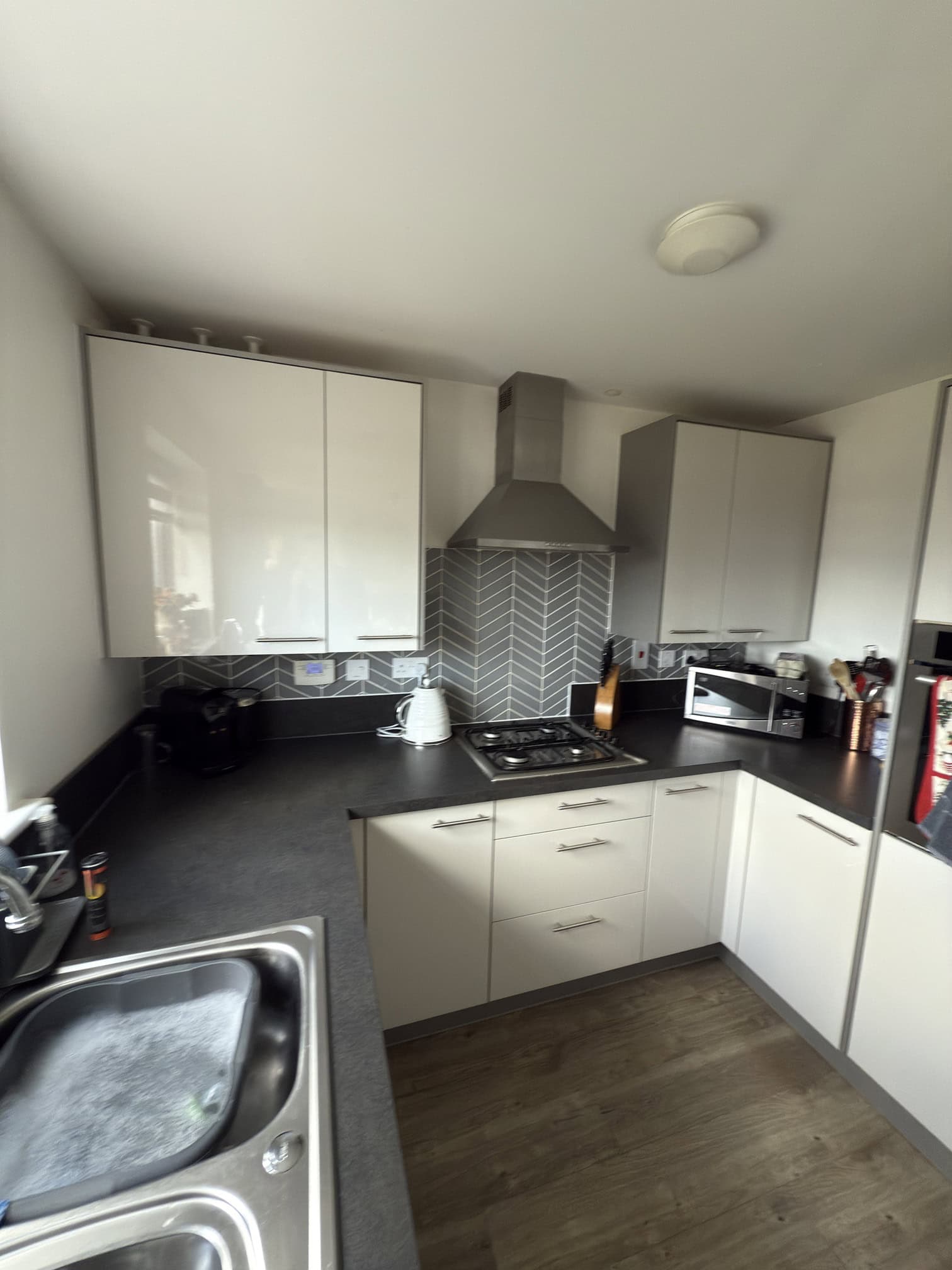 Modern kitchen with white cupboards, a gas cooker with a stainless steel extractor hood, a sink, and various appliances including a microwave, toaster, and kettle. The splashback has a grey chevron pattern, and the floor is wood.