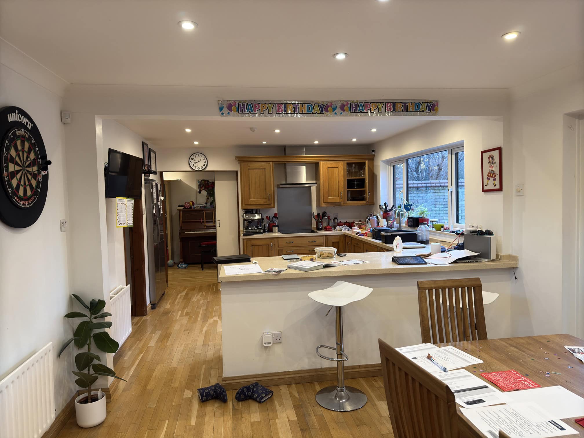 A tidy kitchen and dining area decorated with a Happy Birthday banner. There’s a dartboard on the left wall, papers and stationery on the dining table, and various items on the kitchen worktop. Wooden floors and cupboards throughout.