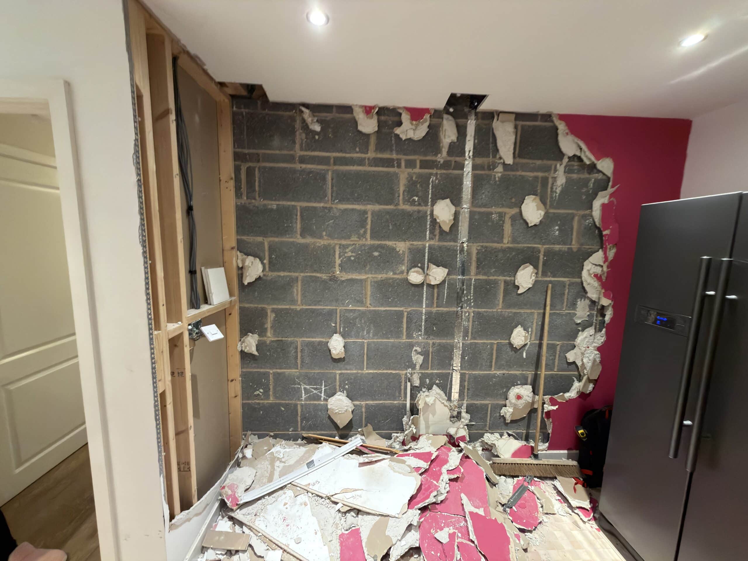 A partially demolished interior wall reveals grey concrete blocks and insulation. Debris, plasterboard pieces, and pink wall fragments cover the floor. A fridge stands to the right, and exposed studs are visible on the left.