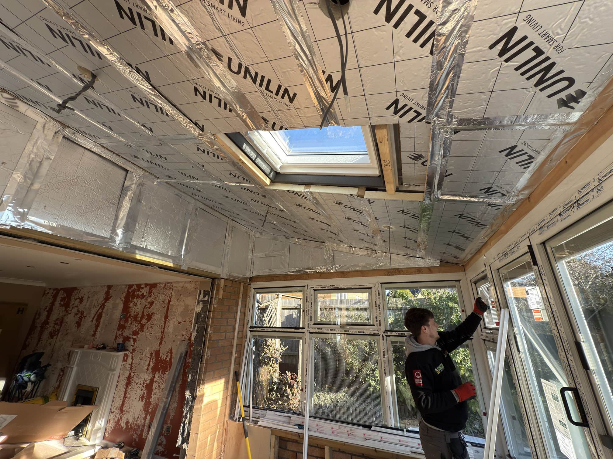 A person fits or repairs windows in a room undergoing renovation, with reflective insulation panels covering the ceiling and part of the walls. Sunlight comes through a skylight and large windows. Tools and materials are scattered inside.