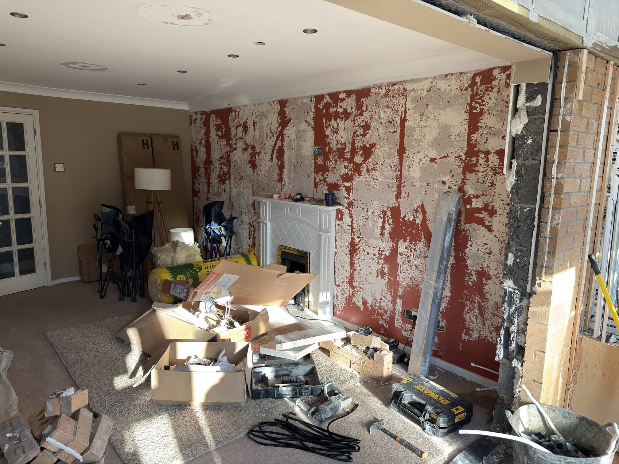 A living room under renovation with stripped red wall, exposed bricks, scattered tools, boxes, and building materials. A white fireplace is on the back wall, and cardboard boxes are stacked in the corner.
