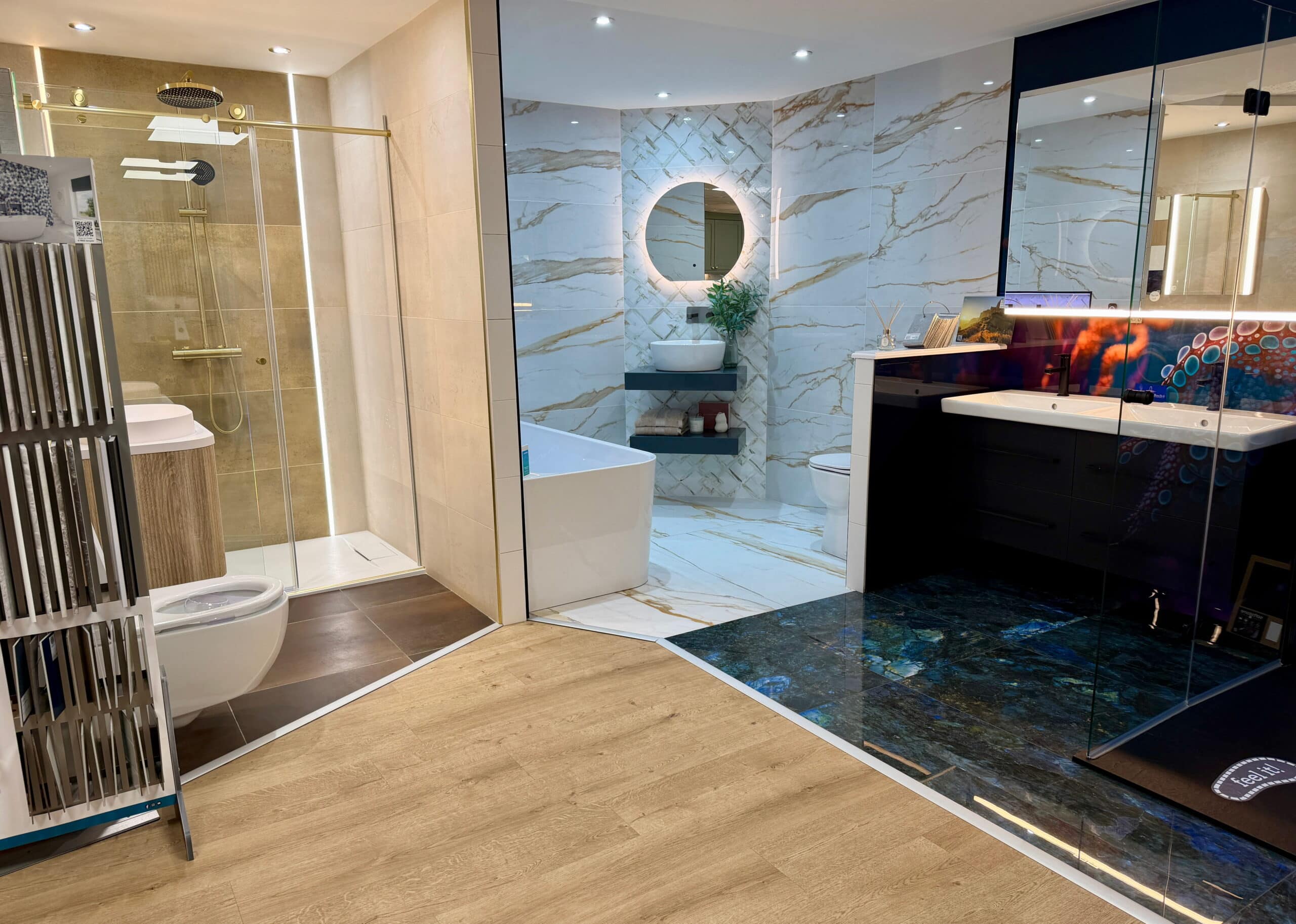 Modern bathroom with glass shower, freestanding bath, black vanity unit, illuminated mirror, and marble walls and floor. Wooden and dark tiled flooring divide the space. Green plant by the bath adds a touch of greenery.