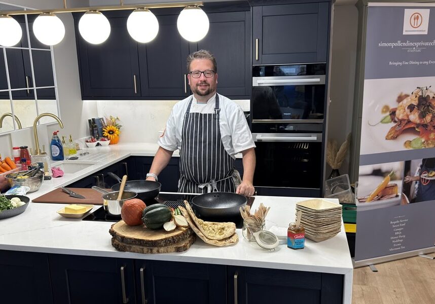A chef in an apron stands in a modern kitchen behind a counter with fresh vegetables, cooking utensils, and crockery—showcasing the sleek design inspired by a leading kitchen and bathroom showroom installation supplier. Dark cupboards and bright lighting set the scene.