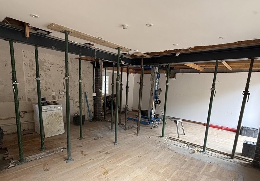 A room under refurbishment with exposed support beams and metal poles holding up the ceiling; brick walls and unfinished surfaces are visible, reflecting a space in progress for kitchen and bathroom showroom installation. Tools and building materials are scattered around.