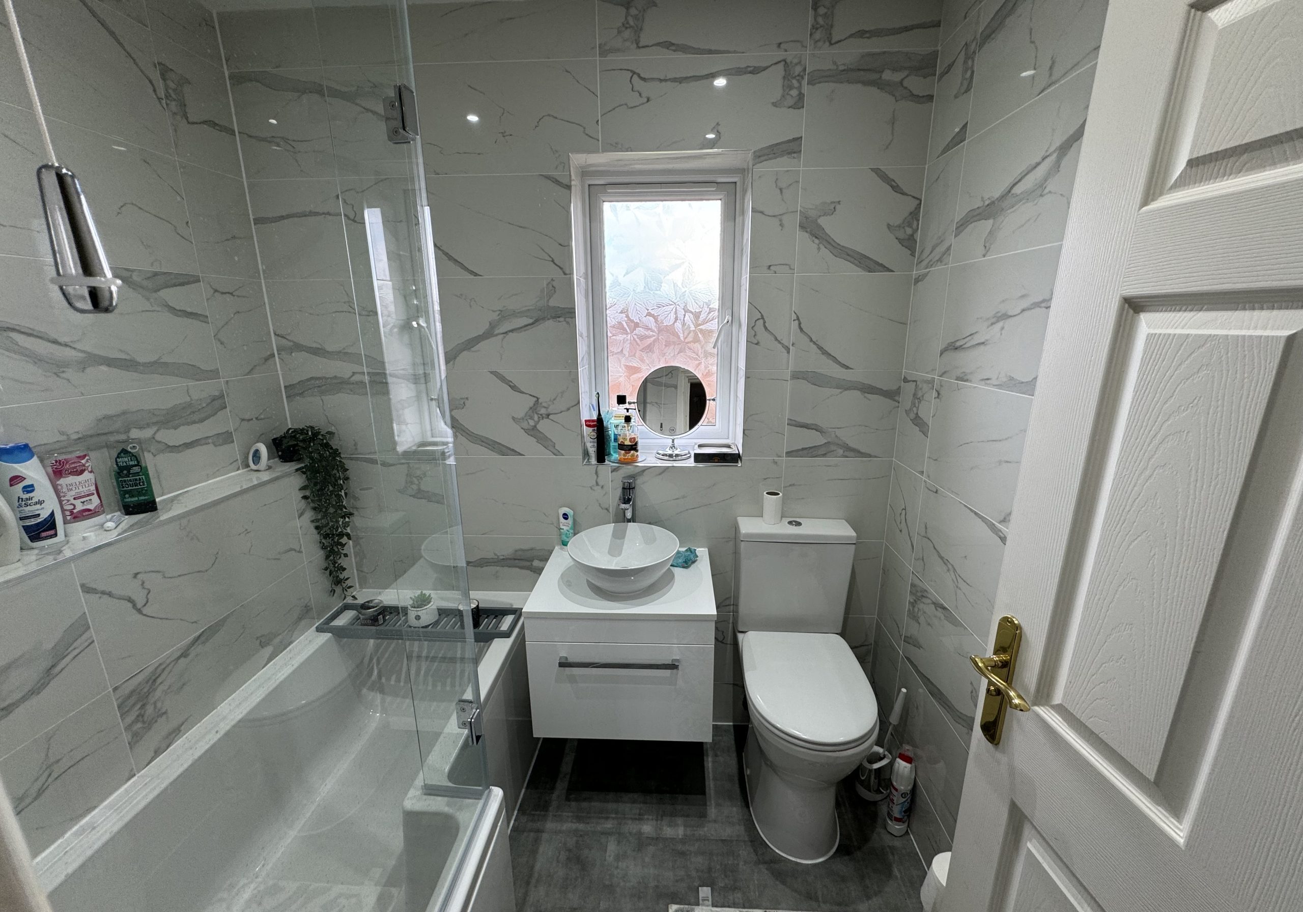 Modern bathroom with grey marble walls, a white bathtub with glass screen on the left, and a stylish vanity—showcasing elegant design perfect for any kitchen and bathroom showroom installation. Door is open, inviting inspiration.